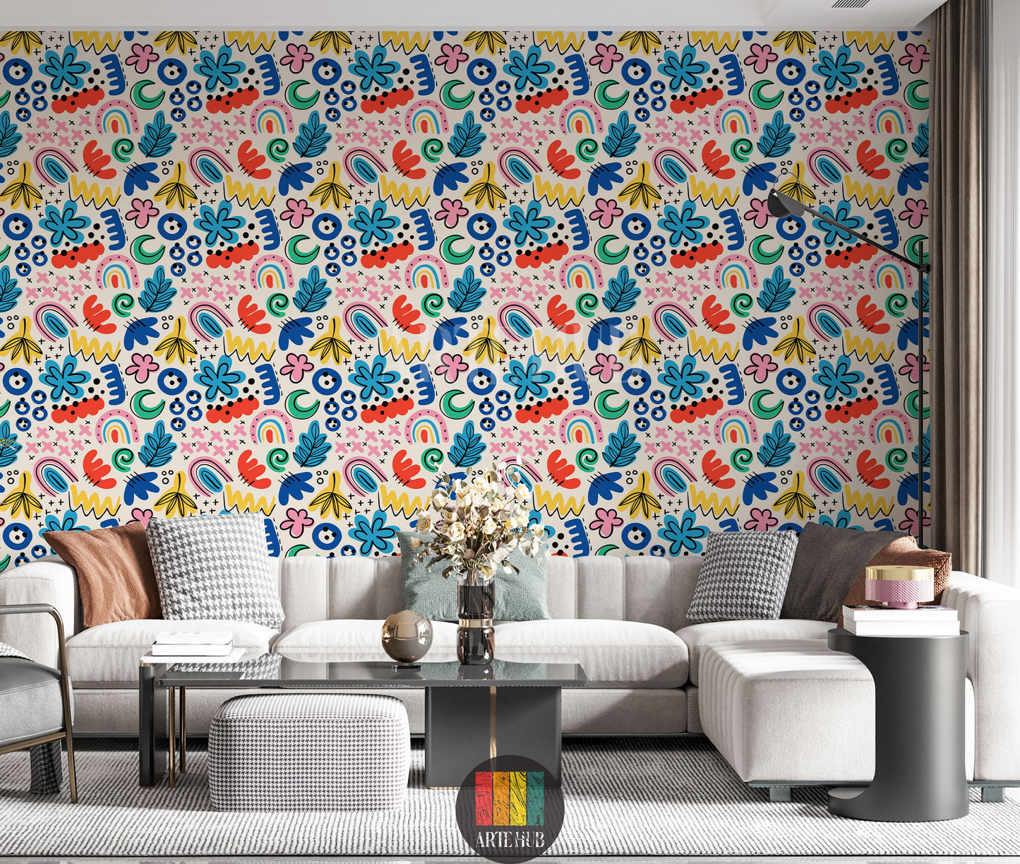  A vibrant living room featuring a wall covered in Whimsical Abstract Colorful Wallpaper. The pattern includes hand-drawn shapes like flowers, leaves, rainbows, squiggles, and dots in bright blues, reds, yellows, pinks, and greens on a light background. A modern grey sectional sofa with various pillows, a black coffee table, and a black floor lamp are visible in the foreground.
 غرفة معيشة مشرقة تتميز بجدار مغطى بورق حائط تجريدي ملون مرح. يظهر النمط رسومات يدوية لأشكال مثل الزهور والأوراق وأقواس قزح و
